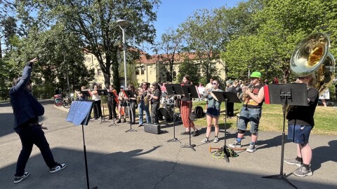 A line of young adults with musical instruments performs outdoors. At the left is a man in dark blue directing the music. 