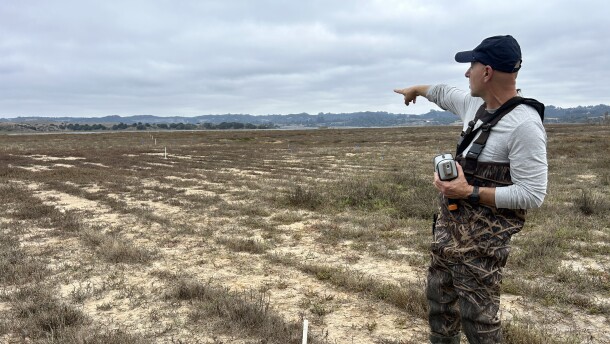 A man wearing boots and waders stands in a marsh field holding a gun-shaped machine in one hand while pointing towards the background, where there's a slough.