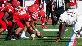 Side view of two high school football teams lined up for the snap of the ball on a green turf field.