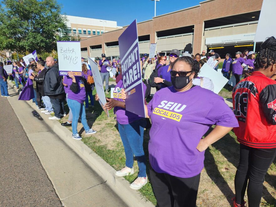 Dozens of Kaiser Permanente workers, many holding purple and white signs, line a sidewalk and grassy area along a road as they strike. A woman in the foreground is wearing a purple union shirt, holding a sign and wearing a black facemask and sunglasses. Others in the background wear similar shirts.