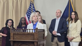 A press conference features a group of individuals standing behind a dark wooden podium bearing the official seal of the State of Michigan Senate.