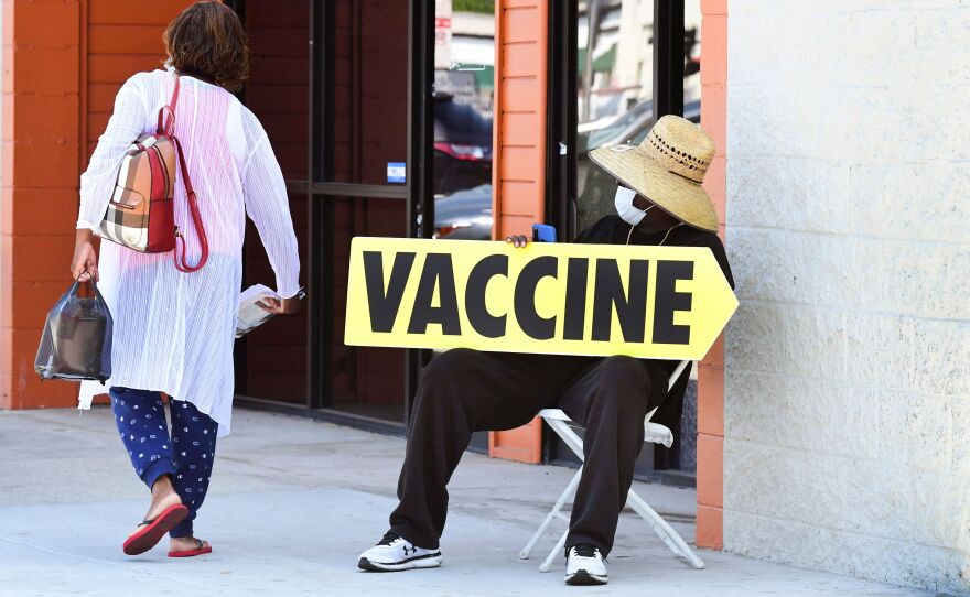 A person sitting down on the sidewalk holds a yellow sign reading "VACCINE" as a woman passes by.