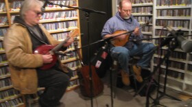 Two men seated on stools in a radio studio playing mandolin