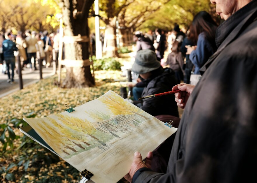 A man paints a watercolor of ginko trees in a crowded park.