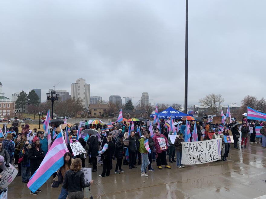 A group of people standing on the steps of the Utah Capitol, many with trans flags and signs. The largest visible sign says "hands off trans lives!"