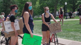 The photo shows several young woman holding signs and wearing face masks. One is also walking a red bicycle.