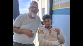 Jonathan Halle sits in a chair with gauze covering both his arms. A male relative with grey hair stands beside him. 