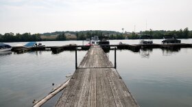 A wooden pier extends into a calm lake with boats docked on either side. The shoreline is lined with trees, and there are gentle hills in the background under a partly cloudy sky.