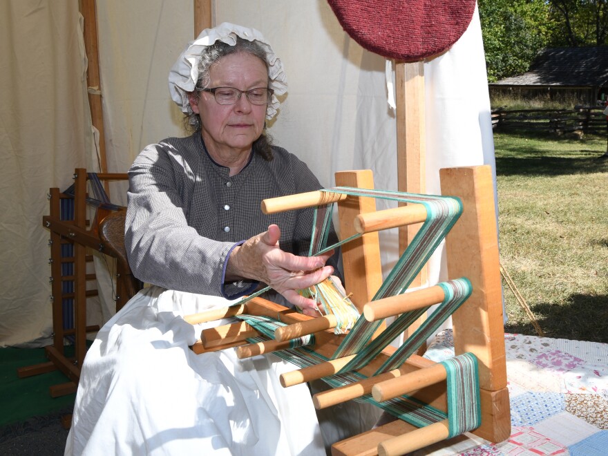 A woman in 1800s period clothing makes thread while sitting in an open-sided tent.