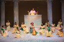 On a stage decorated as a Greek amphitheater, one woman in a Greek costume speaks from a podium to several other women who sit on the steps below. 