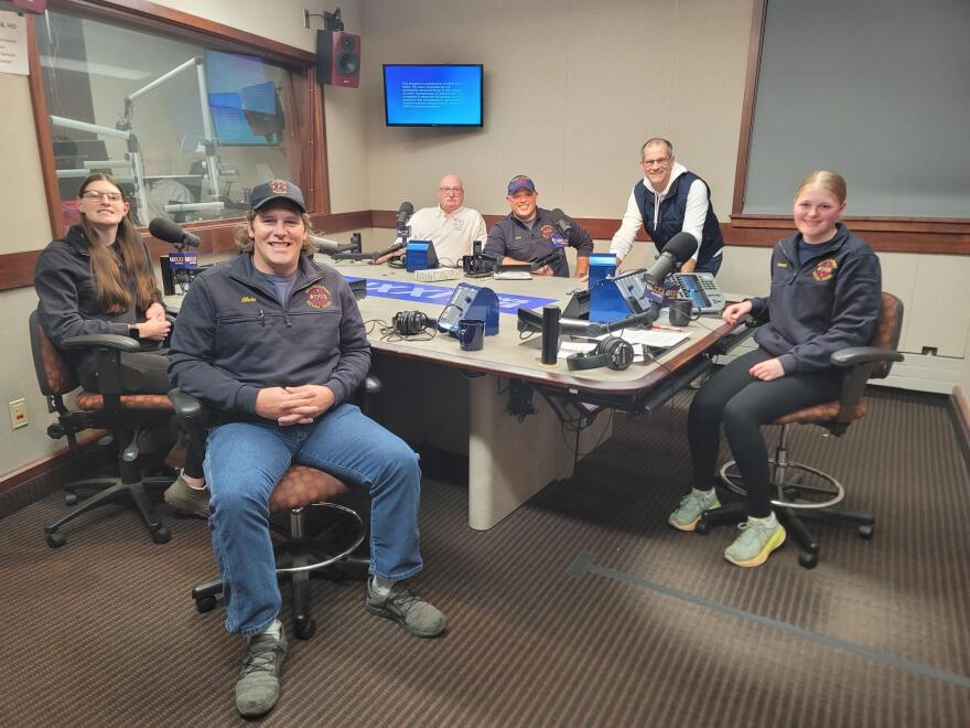 Six people gather around a table in a radio talk studio: a young woman front left has long brown hair and is wearing glasses, a blue fleece pullover and blue pants; a man front right has brown hair and is wearing a blue baseball cap, blue fleece pullover, jeans and grey shoes; a bald man back left has a grey mustache and is wearing glasses and a white polo shirt; a man back center is wearing a blue baseball cap and a blue fleece pullover, a man back right has short dark hair and is wearing glasses, a navy puffy vest, white hooded sweatshirt and blue jeans; a young woman at center wears her blonde hair pulled back and is wearing a blue fleece pullover, black pants and sneakers.
