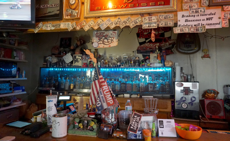 View of a store counter covered in knickknacks and pro-Trump flags. Behind the counter is lots of liquor and more signs, dollar bills, and pro-Trump messaging.