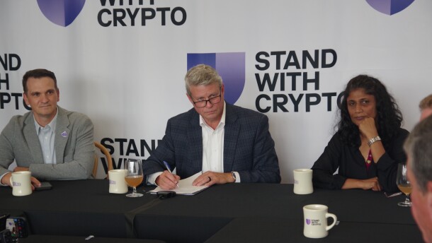 Several people sit around a black table and appear listening to a speaker who is off camera. Behind them, large white banners read "Stand with Crypto"