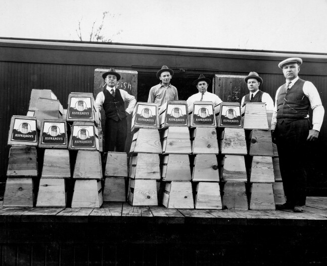 Masuo Yasui (left) stands with four white male produce growers in 1928 (all wearing Fedora hats in a black and white photo) behind crates stacked chest-high with fruits and vegetables. Masuo stands behind two crates stacked knee high.