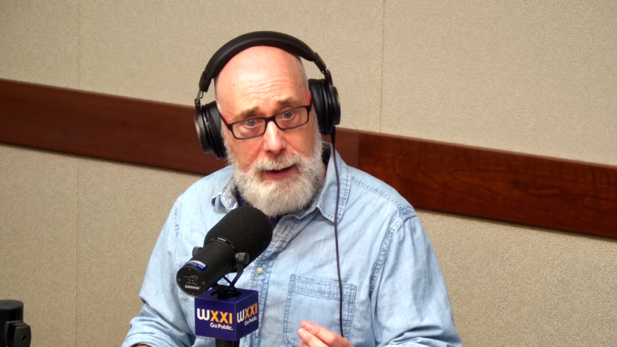 A bald man with a bushy grey beard is sitting in front of a microphone at a table in a radio talk studio and wearing eyeglasses and a light blue button-down shirt.