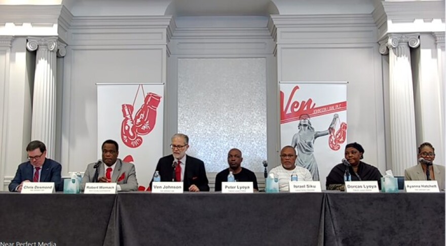 Screenshot showing seven people sitting at a table with microphones in a hotel ballroom.