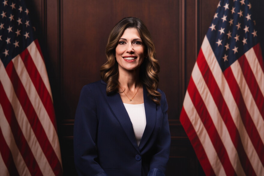 A brunette woman in a navy blue suit smiles in front of an American flag.