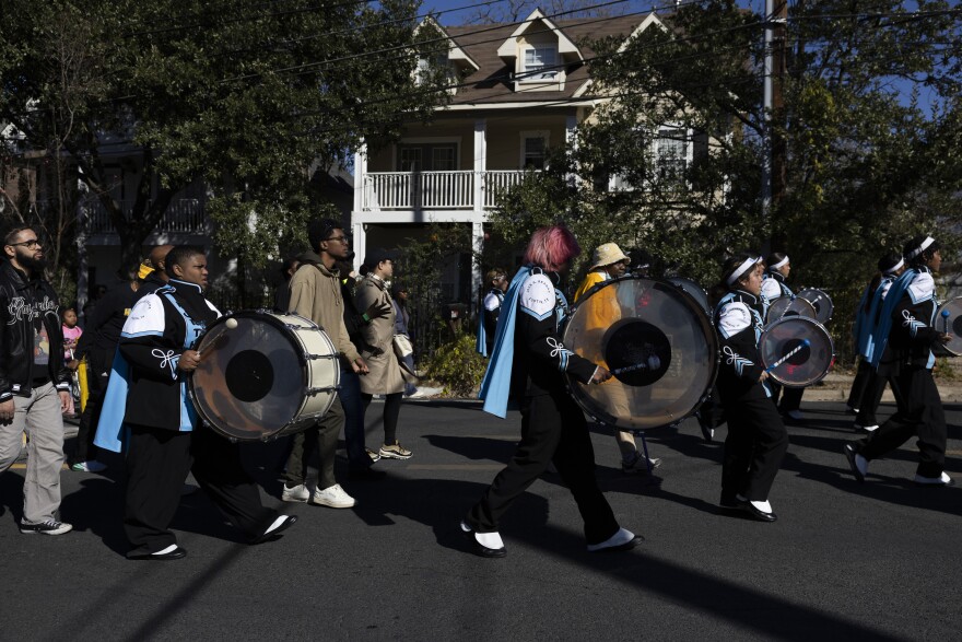 Students in a marching band carrying large drums walk with other people along a street during a celebration honoring Martin Luther King Jr.