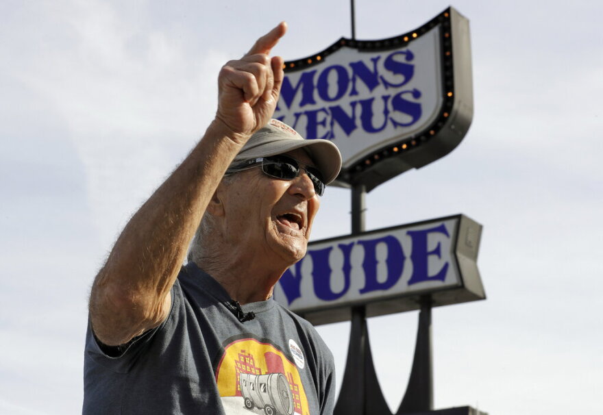 man in sunglasses and cap yells while pointing, standing in front of marquee that reds Mons Venus Nude