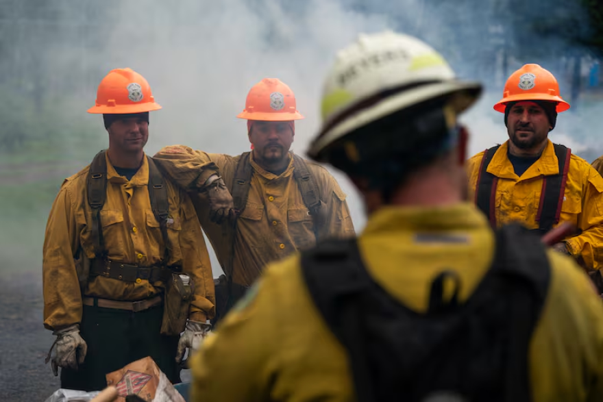 Three people in yellow firefighter gear face the camera while a fourth person has their back to the camera, facing the other three.