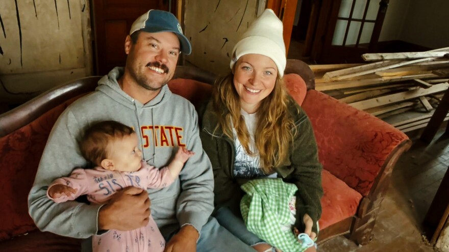 A man and woman sit on a Victorian couch inside an old house that needs restoration. The man is holding a baby.