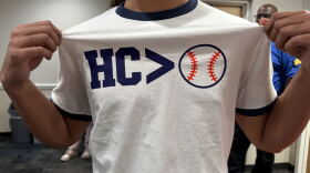 A man grabs the sides of his white shirt to showcase the text that reads "HC > Baseball," but the baseball is an icon, and not words.