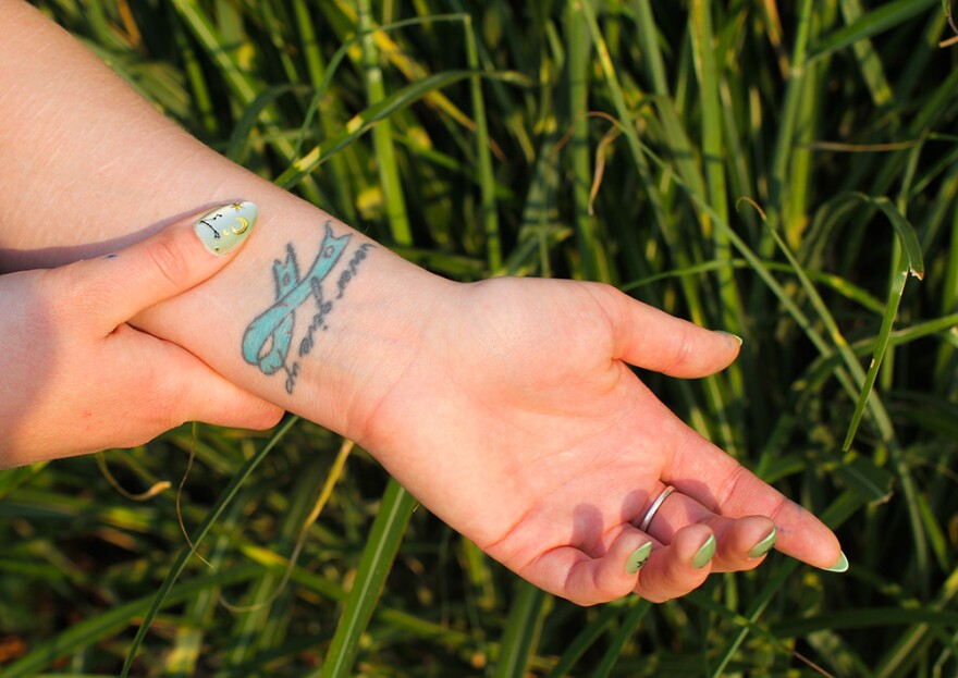 S., a 25-year-old from Clarkston, shows a tattoo she got that says “never give up.” She got the tattoo in 2017 to represent her battle with PTSD, which has affected her mental health for years. The tattered but still-whole blue ribbon represents that even though her experiences left her with trauma, she is still alive. S. says that after surviving her “worst” suicide attempt roughly seven years ago, the tattoo means more to her than ever: “Even though my mind had given up, my body continued to fight for me,” she says. (Credit: Rachel Sun/NWPB)