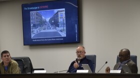 Three men seated at a table with microphones and a screen overhead with a scene of a downtown and the words "The streetscape is defining" on the screen. 