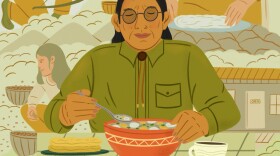 An Indigenous man eats a bowl of soup, surrounded by members of his community sourcing food and cooking it