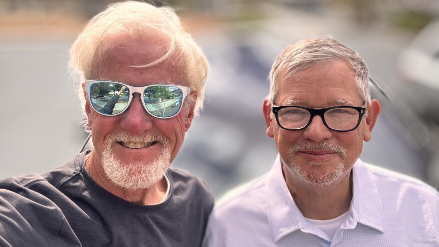 Rich Guy and James Oscarson pose outside James' Tesla in the St. George Sunshine. Each looks a little windblown.  The background is out of focus. Rich stands holding the phone, which can be seen reflected in the white sunglasses he wars. Rich has on a dark grey long-sleeved tee shirt.  He has short white hair and a white goatee.  James is slightly shorter than Rich and wears black framed glassses.  His hair is salt and pepper gray, and he also sports a goatee. James wears a lilac checked button-up shirt with a white tee underneath.