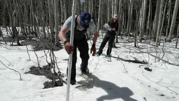 Two people stand next to a metal pole in low snow.