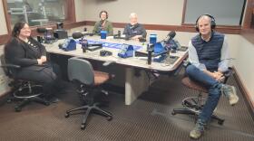 Four people sit at a table in a radio talk studio: a woman front left has long dark hair and is wearing glasses, a black cardigan, black-and-white striped shirt, black pants and black shoes; a man front right has short dark hair and is wearing headphones, a navy puffy vest, white button-down shirt, jeans and brown boots; a man back left has short brown hair, a brown beard and is wearing glasses, a green cardigan sweater and orange shirt; a man back right has short grey hair, a grey beard and is wearing glasses and a black button-down shirt.