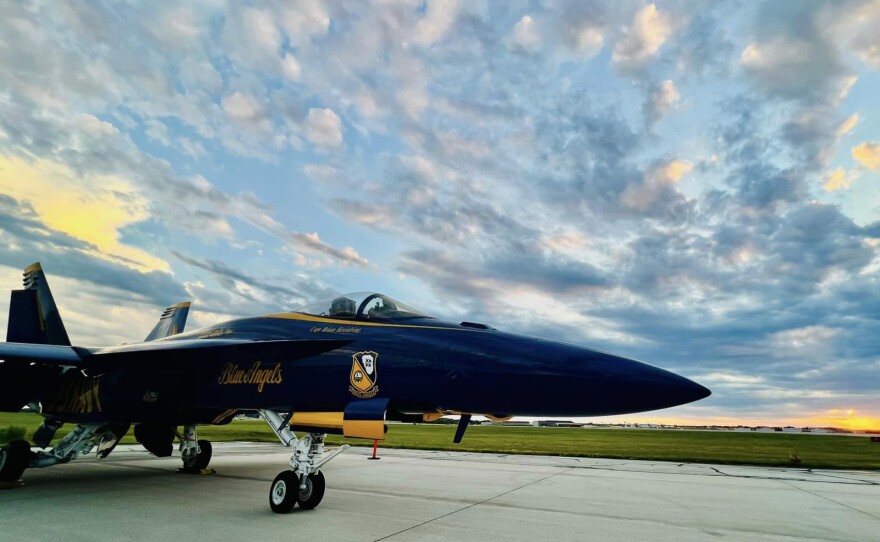 A blue U.S. fighter jet parked on a runway with the sun setting in the background.