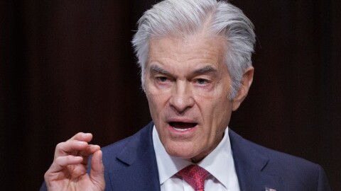 Man with gray hair wearing a blue suit and talking, looking to the left and gesturing with his right hand
