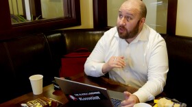 A white man with a brown beard sits in front of his laptop at a desk