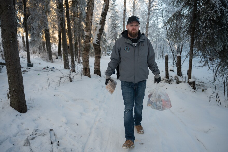 A man walks in a snowy forest carrying a plastic bag of food. 