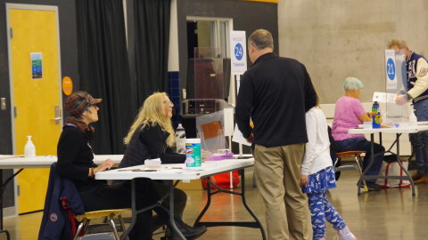 A man in a black shirt and tan pants takes his daughter to a precinct in the Girl Scout headquarters in Kalamazoo.  He's holding his ballot, talking to poll workers as he gets ready to put his completed ballot into the voting machine. 