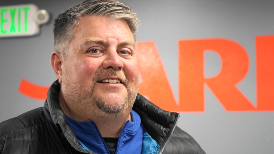 A smiling man stands before a gray-blue wall