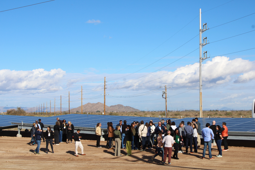 A group of several dozen people stand near an array of solar panels, set up near power lines. There is an arid landscape behind them with mountains in the background.
