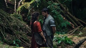 A still from a scene in Hamnet. Jesse Buckley in a red Renaissance dress, stands near Paul Mescal in an English forest. 