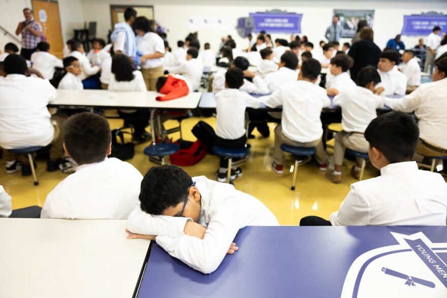 A student is asleep sitting at a cafeteria table. His head rests against his crossed arms. Beside and behind him, other students facing in the opposite direction are sitting at tables and watching a teacher in the background.