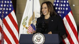 a woman in a suit smiles while standing at a podium with american flags behind her