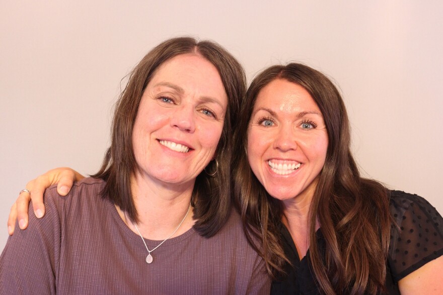 Melissa Julien and Cassie Riding at their St. George StoryCorps appointment on May 10th, 2025. Melissa is taller than Cassie, and leans toward her.  She wears her shoulder length brown hair which she wears parted slightly to her right.  She has on gold hoop earrings and a small silver pendant and chain.  Her tee shirt is plum colored.  Cassie is on Melissa's left side, and has her had on her shoulder in a sideways hug.  Cassie also wears her long dark brown hair parted on her right.  She smiles widely, showing gums.  Her black blouse has translucent, polka-dotted sleeves.