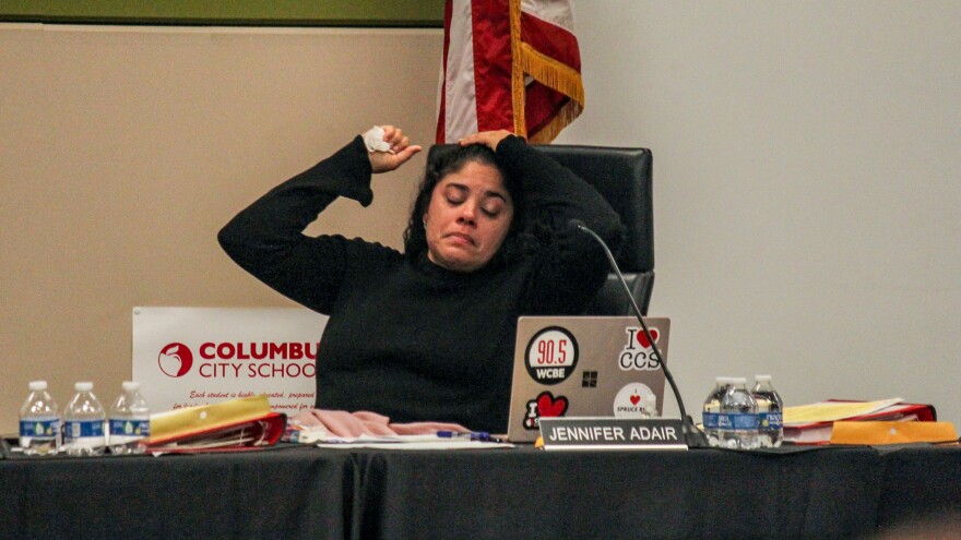 A woman looks distressed as she clutches a tissue and glances down at a laptop. Her name tag reads Jennifer Adair. 