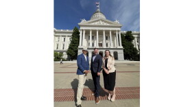 Three people stand in front of the California State Capitol on a sunny day. 