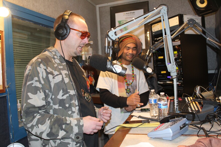 A man with buzzed hair and a trimmed goatee wearing red glasses and a camo jacket over a black t-shirt speaks into a microphone in a radio studio. Another man wearing several hats speaks into another microphone in the background.