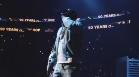 A man in a baseball cap holds a microphone to his mouth while performing on stage in a large arena with an LED ribbon board behind him that reads "50 years of hip-hop."
