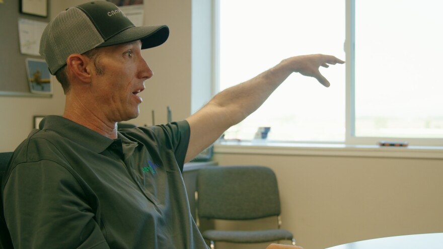 A man in a ballcap and a grey polo shirt, both which read "Crop Air," sits and gestures with his left hand. He's inside an office. Behind him is a window, bulletin board, desk and chair.