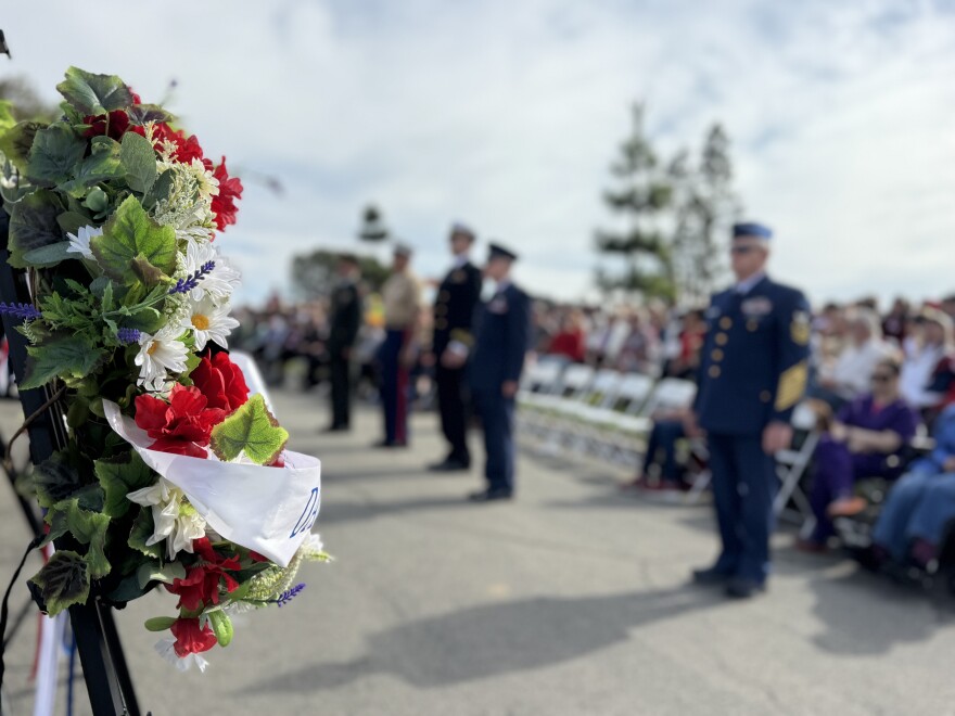 A wreath is positioned in the foreground, with several older men in military uniforms standing at attention behind it.