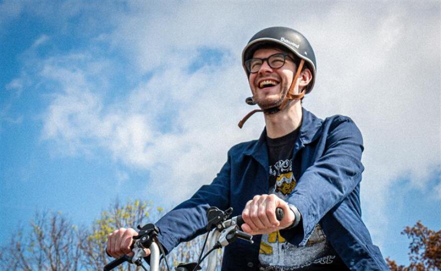 A bearded man wearing a helmet, glasses and a blue jackets rides a bicycle outside.